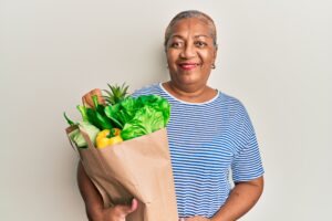 Senior african american woman holding paper bag with bread and groceries looking positive and happy standing and smiling with a confident smile showing teeth